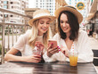 © halayalex - Two young beautiful smiling hipster female in trendy summer white dress and hats. Sexy carefree women drinking fresh vegetable cocktail smoothie drink in plastic cup with straw. Using phone apps
