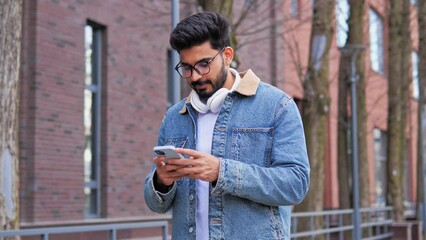 Good looking Latino man with headphones walks down the street, chatting on his mobile phone, typing a message while scrolling. Surfing social media, enjoying his free time and the holiday concept.