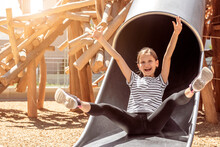 Happy Child Sliding Down Slide Having Fun On Children Playground. Joyful Kid Girl Enjoying Playing Outdoors. Childhood Active LLeisure Summer Holiday.