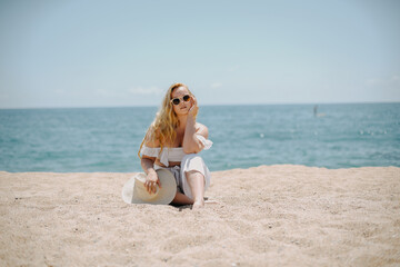 Wall Mural - Happy girl in white outfit sits on sand against the backdrop of the sea or ocean beach. Woman smiles and laughs, vacation and joy. Fashion model, beautiful jewelry, earrings, hat, sun glasses