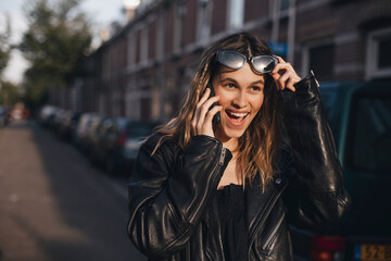 Wall Mural - Attractive happy woman talking on cell phone, raised her glasses in surprise, open mouth. Young cute tourist blonde woman with two thin braids outside, wearing black leather jacket and black dress.