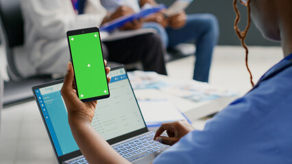 African american woman holding smartphone with greenscreen, using chroma key display in hospital reception. Person looking at isolated copyspace with mockup background, waiting room.