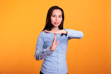 Filipino woman showing timeout and break gesture in studio, doing t shape sign with hands to pause action. Serious young adult gesticulating refuse standing over yellow background. Stop concept