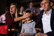 Young woman looking shocked while watching movie in cinema, closing eyes of her preteen son with her hand. Concept of parental guidance and protection