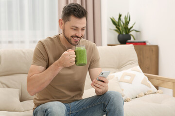 Poster - Happy man holding delicious fresh smoothie and using smartphone at home