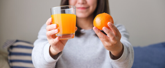 Wall Mural - Closeup image of a young woman holding and showing an orange and a glass of fresh orange juice at home