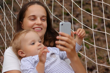 Wall Mural - Portrait of young smiling attractive woman with her daughter, mother and child relaxing in hammock in forest and using smart phone, looking at device screen, watching online cartoons together.