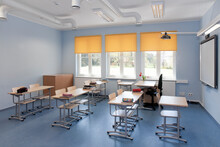 A School Classroom With Desks And Chairs And Yellow Window Blinds.