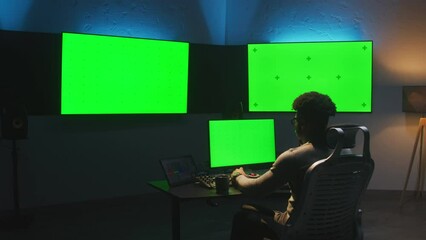 Fototapete - African American man works on computer in studio with equipment. Multiple chroma key big screen on the wall. Professional software displayed on tablet. Color grading or film editing room. Dolly shot.