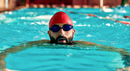 Swimming - male swimmer swimming breaststroke. Close up portrait of man doing breast stroke swimming in pool wearing red swimming cap and swim goggles