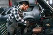 © standret - Mechanic is using a tool to repair the car under the hood. Man in uniform is working in the auto salon
