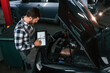 © standret - Standing near the car with hood opened. Holding tablet. Man in uniform is working in the auto salon