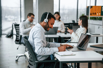 Man is sitting by laptop in headphones. Four people are working in the office together