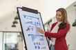 © ArLawKa - Asian businesswoman in suit standing smiling holding pen pointing at document chart graph on whiteboard presenting data in conference room