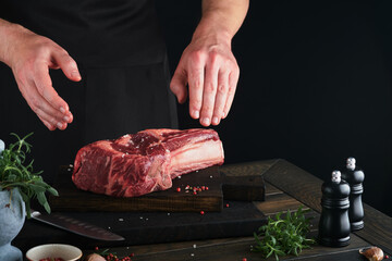 Wall Mural - Chef cutting steak beef. Mans hands hold raw steak Tomahawk on rustic wooden cutting board on black background. Cooking, recipes and eating concept. Selective focus.