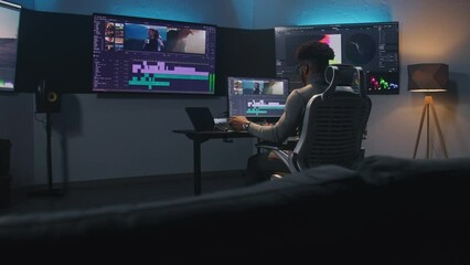 Fototapete - African American multimedia artist drinks coffee and makes color grading in editing program on computer. Process of video post production in modern studio. Multiple monitors with travel movie footage.