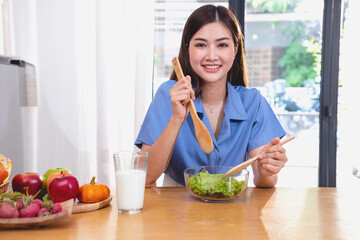 A young woman with a beautiful face in a blue shirt with long hair eating fruit sitting inside the kitchen at home with a laptop and notebook for relaxation, Concept Vacation.