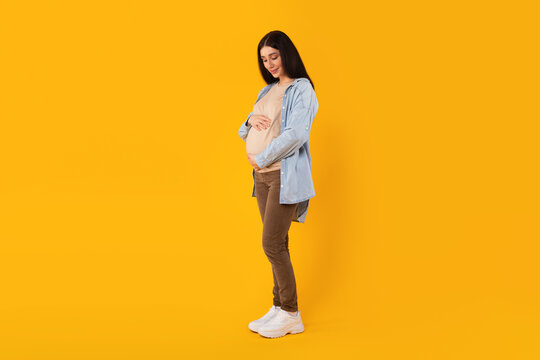 Wall Mural - Full body length shot of pregnant lady keeping hands on big belly isolated on yellow studio background, free space
