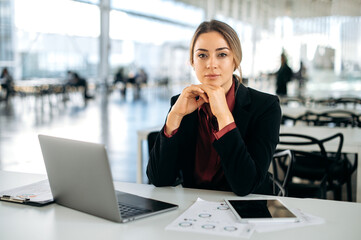 Beautiful confident successful influential caucasian business lady, in elegant clothe, director of the company, sits at a work desk in the office, looks at the camera with a slight smile