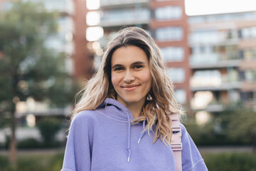 Wall Mural - Close up portrait attractive young happy woman with fresh and clean skin stands outside city near building. Smiling curly blonde wear purple hoody and look at camera. Lifestyle, female beauty concept.