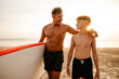 © Maskot - Smiling man holding paddleboard with arm around son at beach during sunset