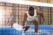 View through net of concentrated sporty african american man playing paddle tennis on indoor court, preparing to hit forehand