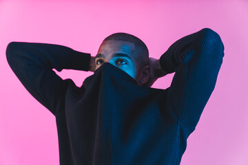 Cool and fashionable man. Young Black guy hiding his face in his sweatshirt and holding hands behind his neck. Studio pink background. High quality photo