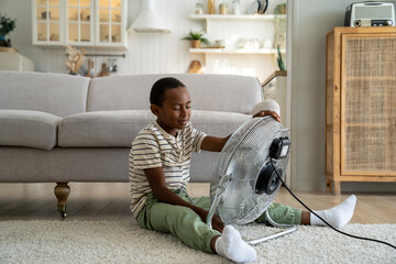 Wall Mural - Small African American kid sitting alone in living room enjoy fresh cool air blowing from electric fan. Little kid suffering from heat at home, cooling down during hot weather. Children and heatwave