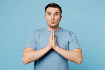 Wall Mural - Young smiling happy caucasian man wear casual t-shirt hold hands folded in prayer gesture, beg about something isolated on plain pastel light blue cyan background studio portrait. Lifestyle concept.