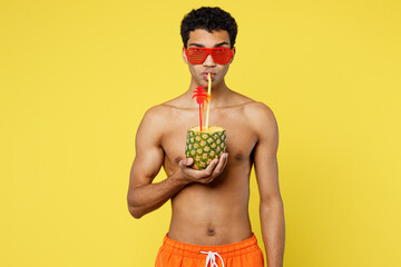 Young handsome sexy man wearing orange shorts swimsuit glasses drink pineapple cocktail look aside relax near hotel pool isolated on plain yellow background. Summer vacation sea rest sun tan concept.