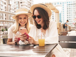 Wall Mural - Two young beautiful smiling hipster female in trendy summer white dress and hats. Sexy carefree women drinking fresh vegetable cocktail smoothie drink in plastic cup with straw. Using phone apps