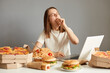 © sementsova321 - Image of sleepy tired woman working or watching movie on laptop during long hours isolated over gray background, being surrounded with pizza, pasta, hotdogs, hamburgers.