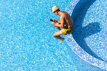 High Angle View Of Young Man Using Smartphone In Resort Hotel Poolside During Summer Holidays, Vacation. Guy Hold Mobile Phone Isolated On Blue Water Swimming Pool Background Copy Space.