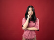© Daniel - A portrait of a young Indonesian (Asian) woman wearing a pink shirt, looking puzzled with one hand folded and the other on her chin. Isolated with a red background