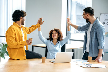 Cheerful colleagues, multiracial team, sitting in a creative office, working on a new project, a startup, developing a strategy and deadlines, happy with the result, celebrate success, achievement