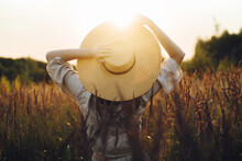 Stylish Boho Woman With Straw Hat Posing Among Wild Grasses In Sunset Light, Back View. Summer Travel. Young Carefree Female In Rustic Linen Cloth Relaxing In Summer Meadow. Atmospheric Moment