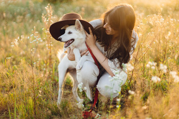 Stylish happy woman playing with cute dog with hat among wildflowers in sunset light. Summer travel with pet. Young carefree female having fun with white danish spitz in summer meadow