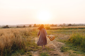 Wall Mural - Stylish woman with straw hat dancing at oat field in sunset light. Atmospheric happy moment. Young female in rustic linen dress relaxing in evening summer countryside, rural life