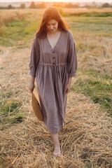 Poster - Stylish woman with straw hat walking at oat field in sunset light. Atmospheric tranquil moment. Young female in rustic linen dress relaxing in evening summer countryside, rural slow life