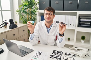 Sticker - Young hispanic oculist holding glasses and contact lenses smiling looking to the side and staring away thinking.