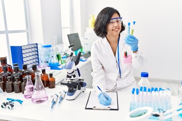 Poster - Young latin woman wearing scientist uniform holding test tube writing on clipboard at laboratory