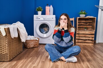 Poster - Young hispanic girl doing laundry shocked covering mouth with hands for mistake. secret concept.