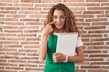 Sticker - Young caucasian woman holding art notebook pointing with hand finger to face and nose, smiling cheerful. beauty concept