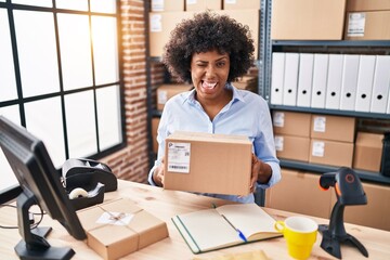Wall Mural - Black woman with curly hair working at small business ecommerce holding box sticking tongue out happy with funny expression.