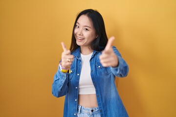 Canvas Print - Young asian woman standing over yellow background pointing fingers to camera with happy and funny face. good energy and vibes.