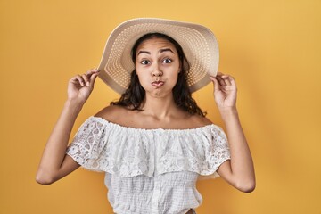 Poster - Young brazilian woman wearing summer hat over yellow background puffing cheeks with funny face. mouth inflated with air, catching air.