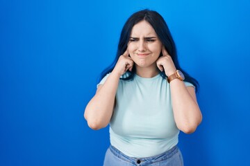 Canvas Print - Young modern girl with blue hair standing over blue background covering ears with fingers with annoyed expression for the noise of loud music. deaf concept.