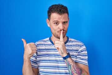 Sticker - Young hispanic man standing over blue background asking to be quiet with finger on lips pointing with hand to the side. silence and secret concept.