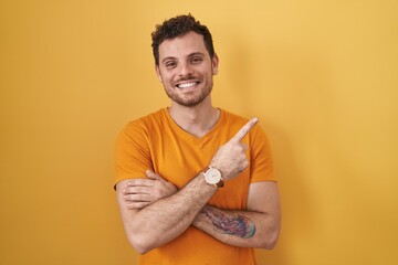 Wall Mural - Young hispanic man standing over yellow background with a big smile on face, pointing with hand finger to the side looking at the camera.