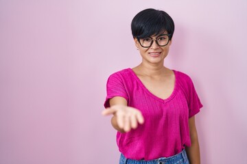 Sticker - Young asian woman with short hair standing over pink background smiling cheerful offering palm hand giving assistance and acceptance.